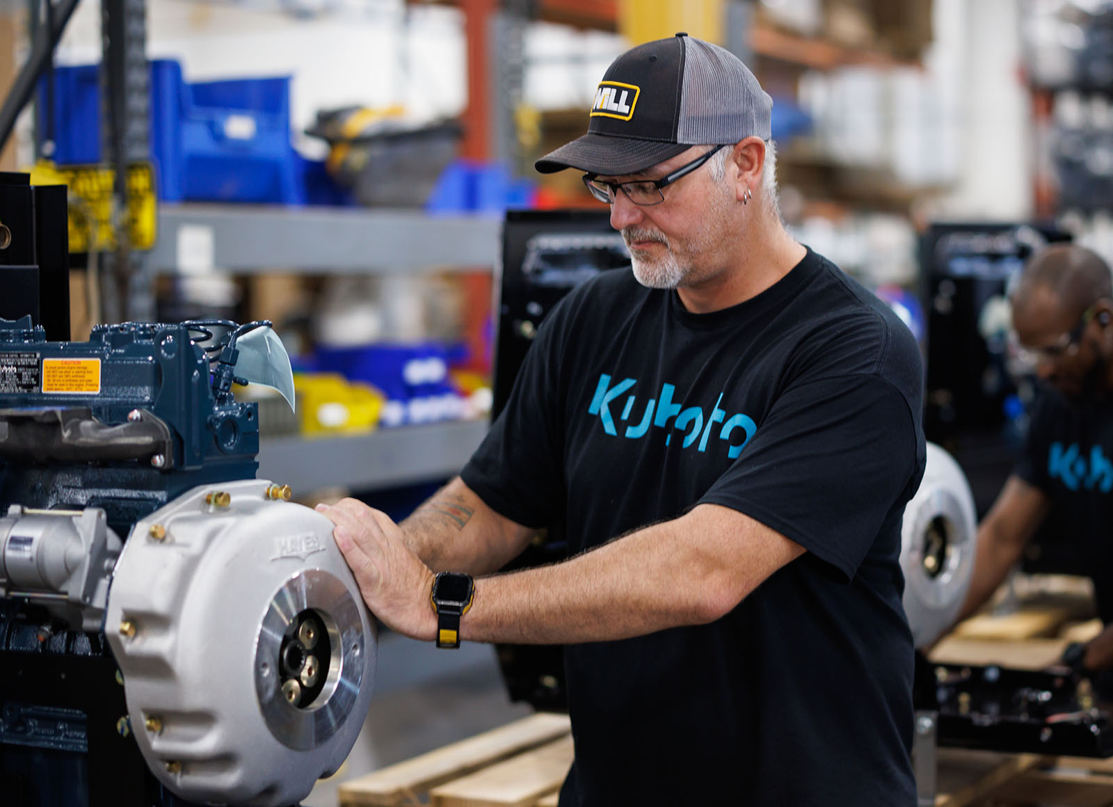 A man wearing a Kubota shirt is crouched over, performing maintenance on a engine.